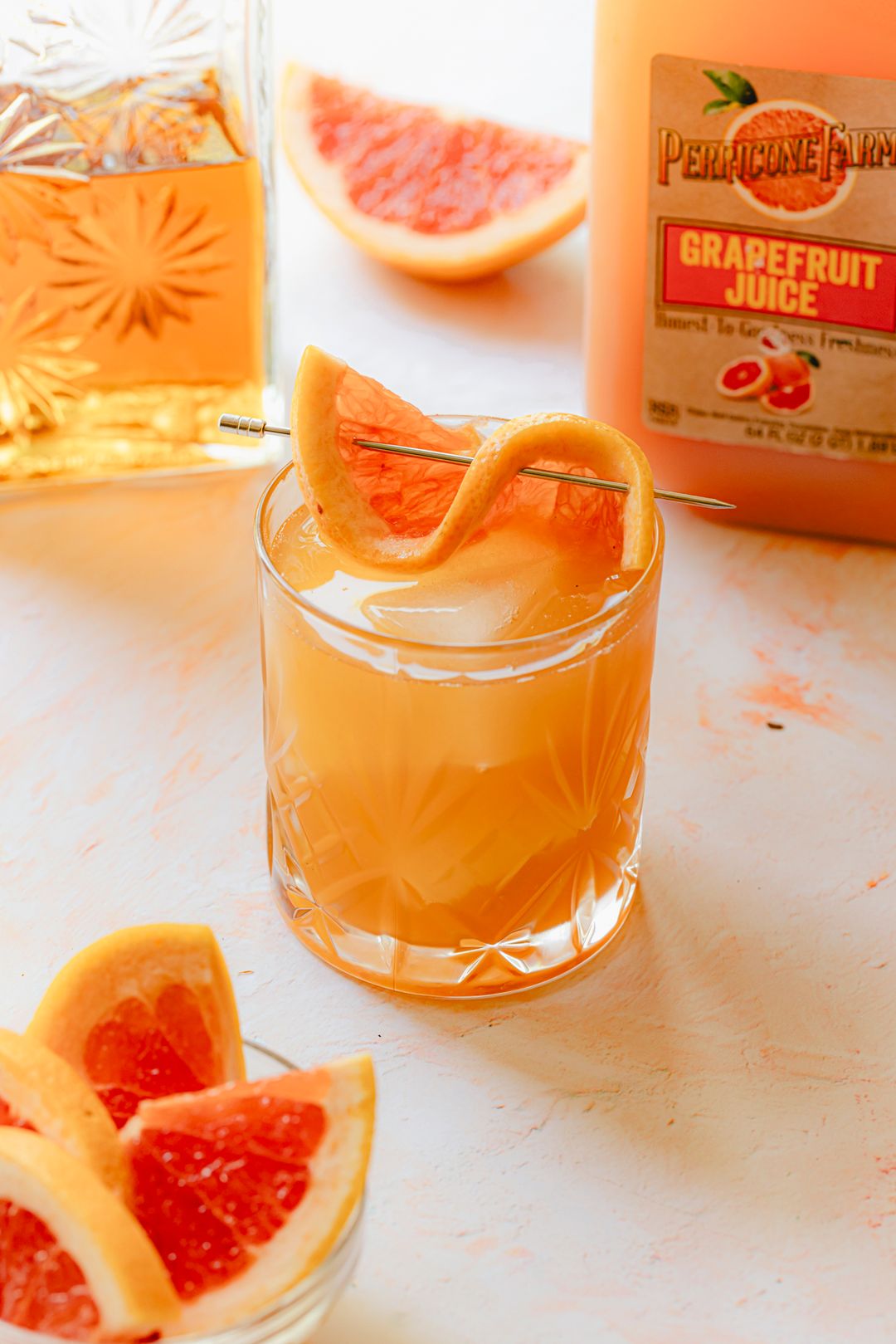 Smokey Grapefruit Old Fashioned made with Perricone Farms Grapefruit Juice, bourbon, and maple syrup, garnished with grapefruit peel beside a bottle of juice and sliced citrus
