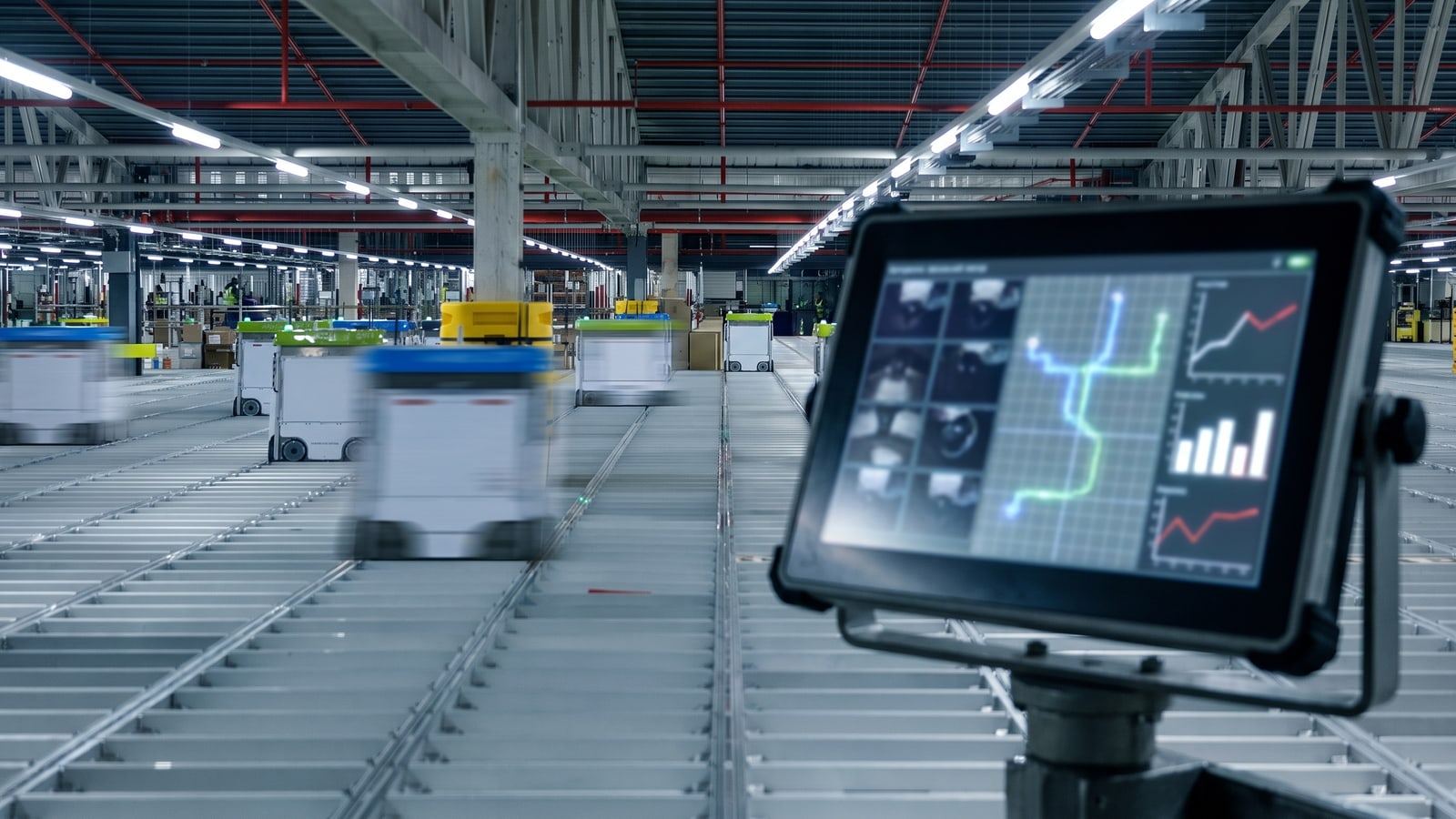 Real boxy warehouse robots running on a metal grid in an automated fulfilment centre, with an out-of-focus operations screen showing fleet telemetry.