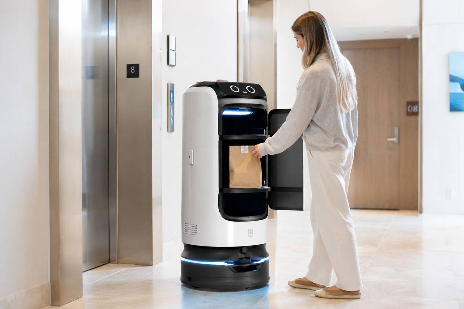 A hotel guest in a softly lit hallway takes a paper bag from the open compartment of a white Pudu delivery robot waiting by the elevator — robotics as a working, revenue-generating service.
