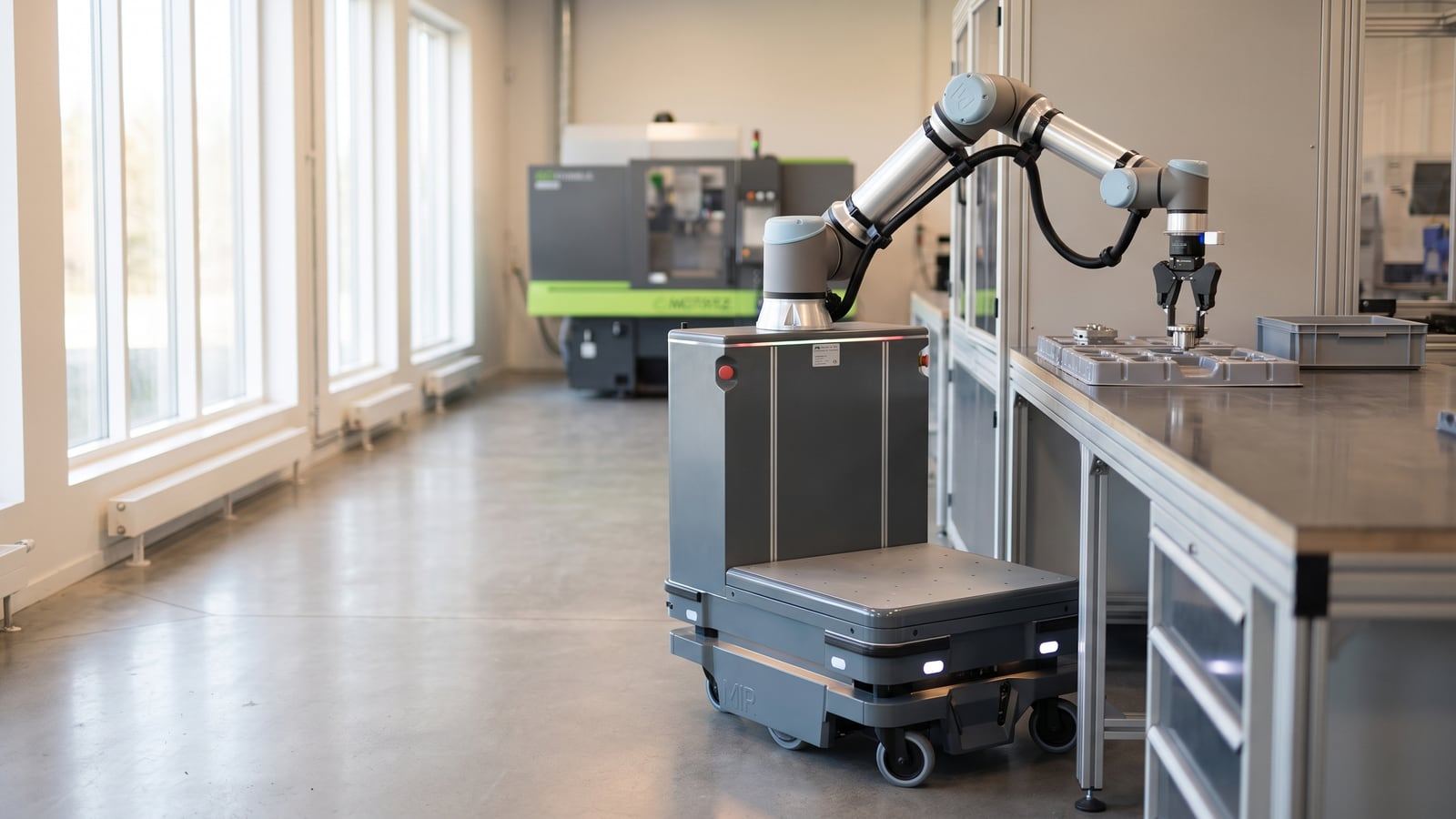 A real mobile manipulator robot with a grey wheeled base and a collaborative arm picks a small component from a tray at a workbench in a bright production facility.