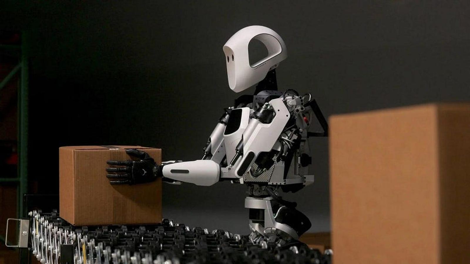 Ein weisser humanoider Roboter hebt einen Karton von einem Rollenförderband an einer Lager-Paketstrasse.