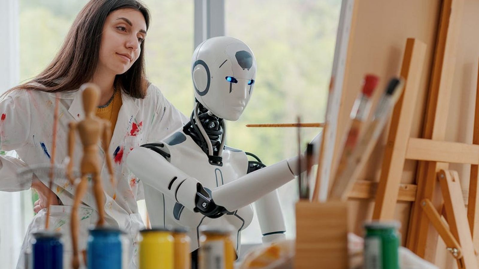 Ein humanoider Roboter malt an einer Staffelei neben einer jungen Frau im farbbespritzten Kittel in einem sonnigen Atelier.