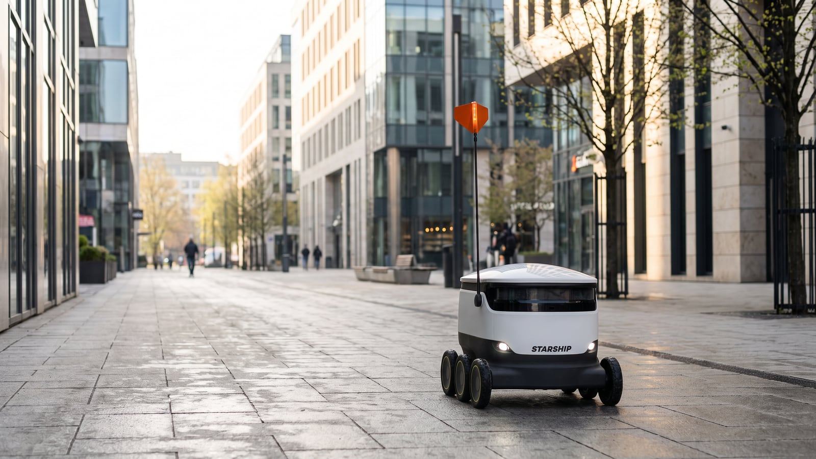 A real six-wheeled white delivery robot drives along a sidewalk in a bright modern European city district on a delivery run, soft morning daylight.