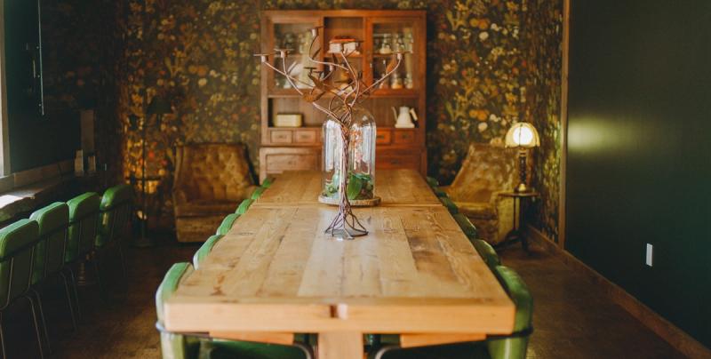 A long wooden communal table with green chairs in a dim lobby lounge, a glass-fronted hutch against a stone wall.