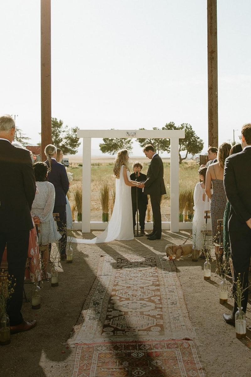 An outdoor wedding ceremony: a couple at a white arch facing open grassland, guests standing either side of a rug-lined aisle.