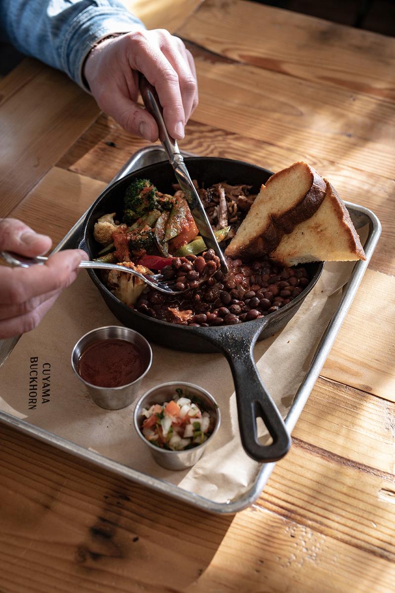 A cast-iron skillet of stewed beans and greens with toast, being served at a wooden table.