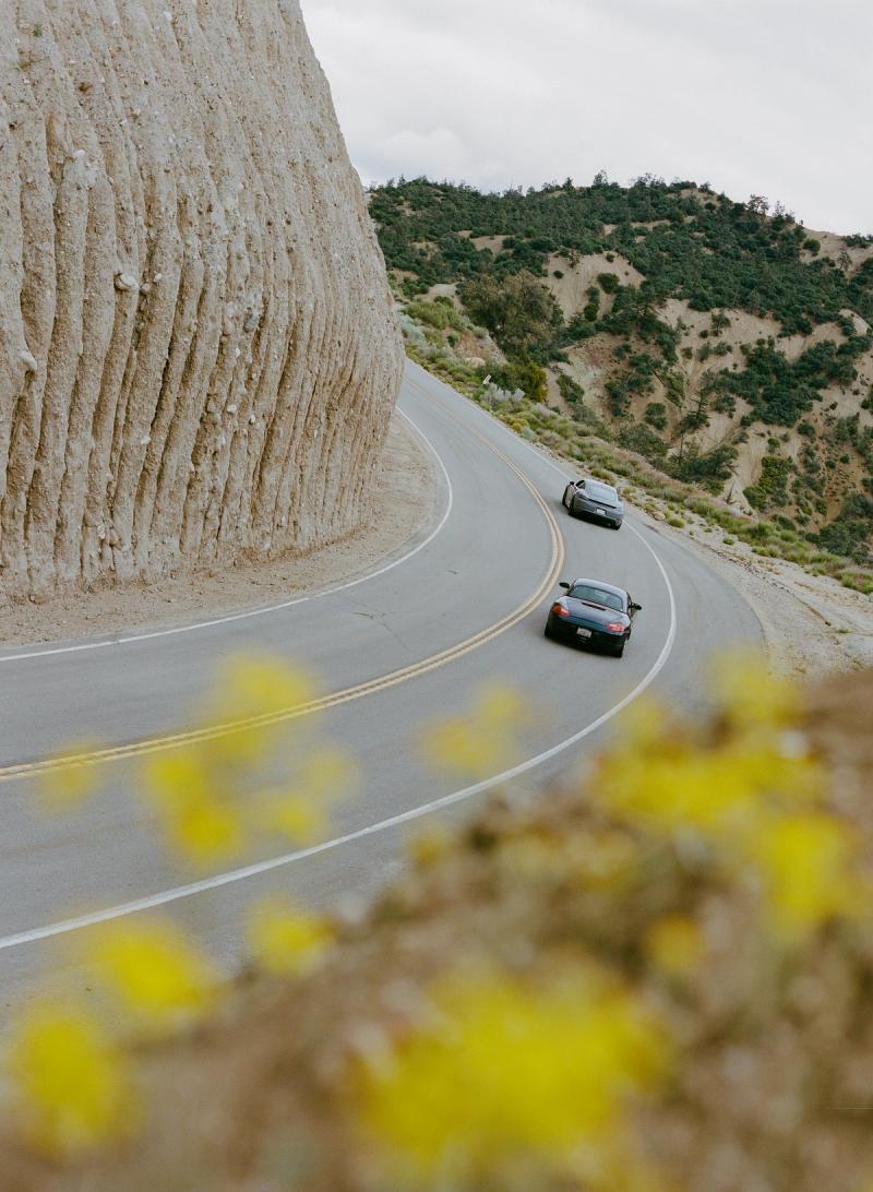 Two cars rounding a bend on Highway 33 beneath a sandstone cliff, wild mustard in the foreground.