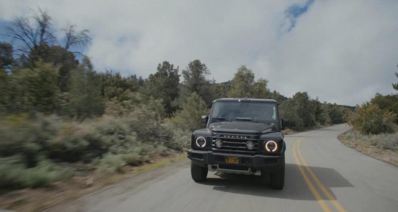 A dark SUV driving a winding two-lane mountain road lined with brush and pines.