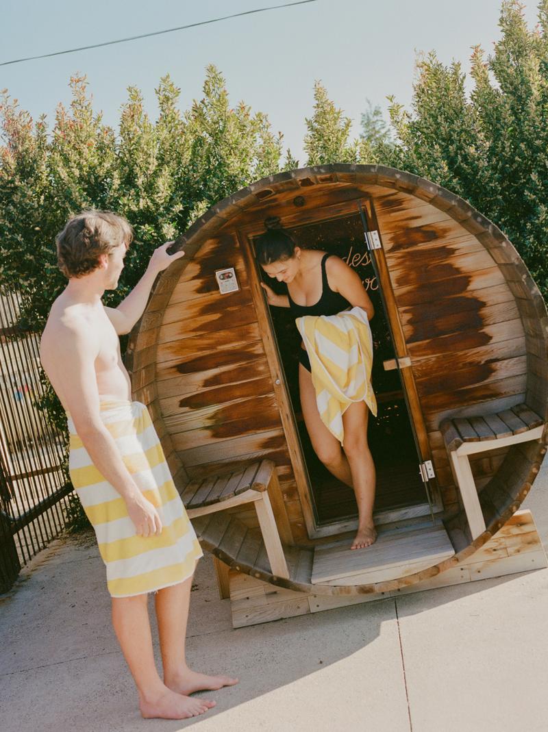 Two guests in robes stepping into the barrel sauna beside the pool.