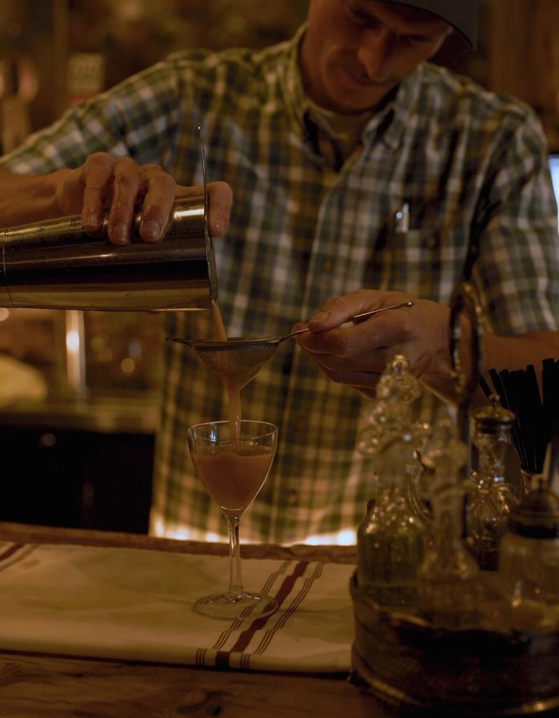 Bartender in a plaid shirt pouring a cocktail from a shaker into a coupe glass