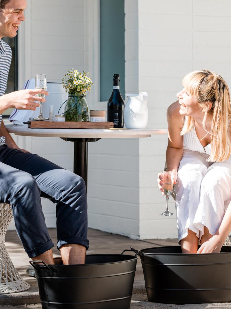 Two guests soaking their feet in galvanised tubs outside a whitewashed cabin.