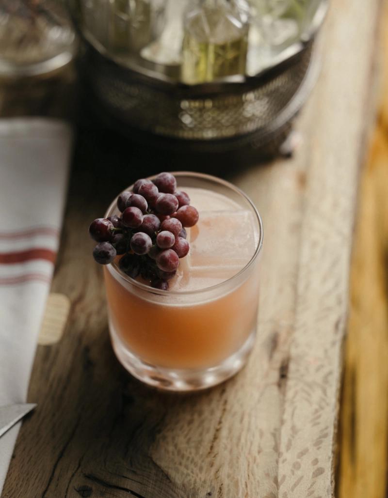 A pale pink cocktail over ice on a wooden tray, garnished with a cluster of frosted grapes.