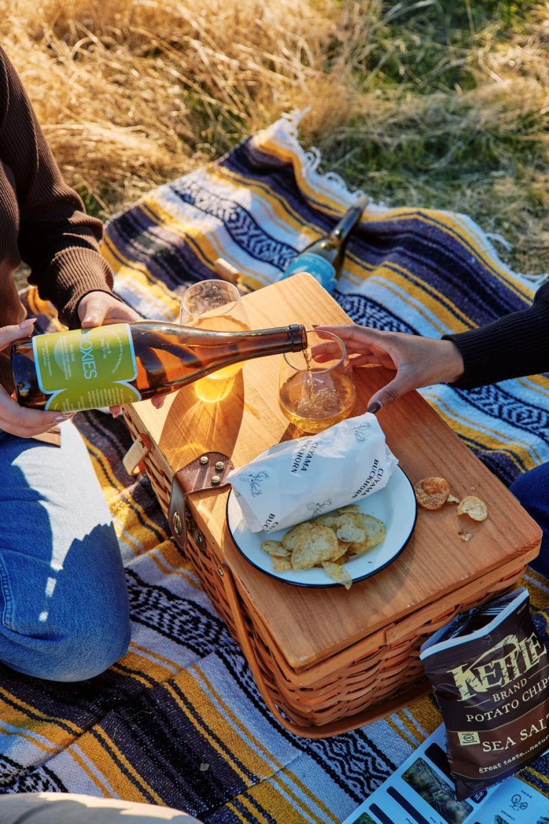 Hands pouring a drink beside a picnic basket of chips and snacks on a blanket