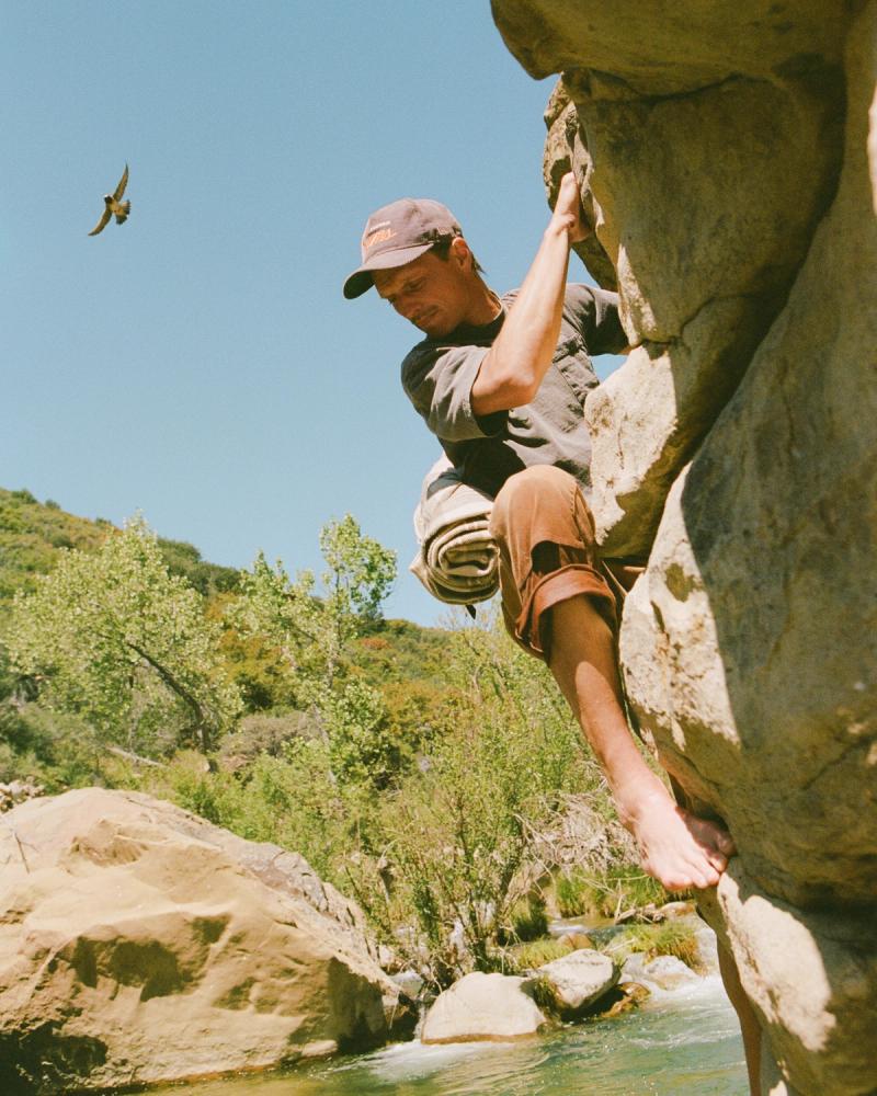 A climber reaching for a hold on a sandstone boulder above a creek, a hawk in the sky behind.
