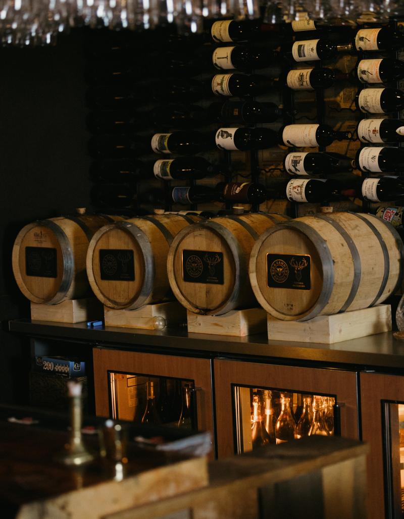 Small oak barrels on the back bar, in front of a wall of racked wine bottles.
