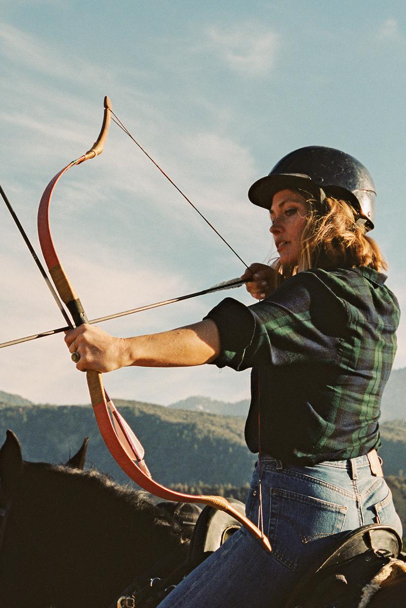 A woman in a green plaid shirt drawing a bow on horseback, with mountains behind her.