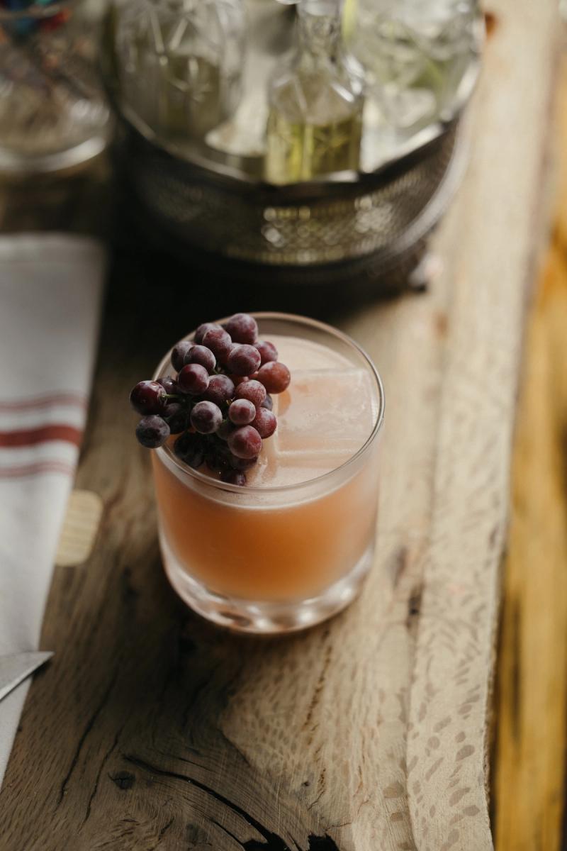 A blush-coloured cocktail garnished with grapes, resting on a weathered wooden bar.