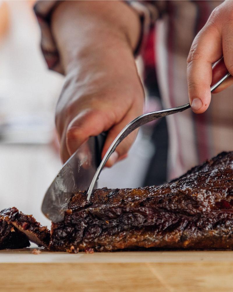 A cook slicing a smoked brisket with a carving knife over a wooden board.