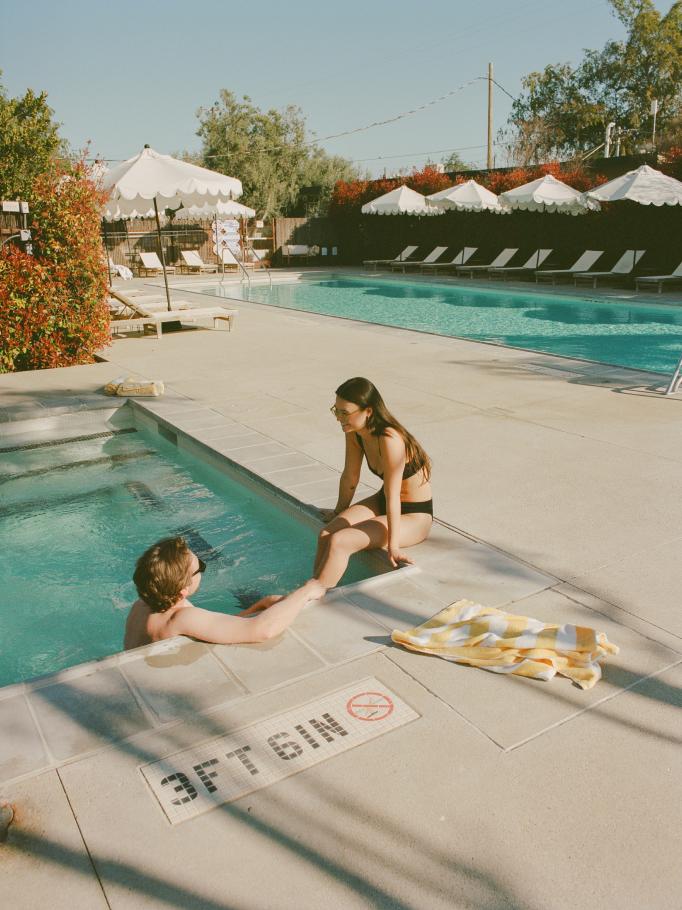Two friends chatting at the pool's edge beside striped umbrellas