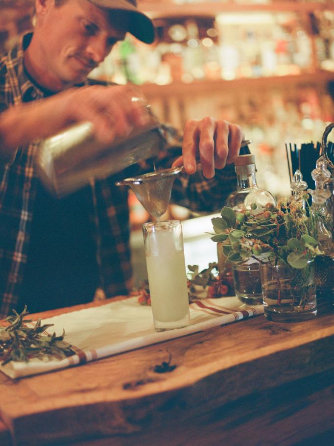 A bartender shaking a cocktail through a strainer over fresh herbs