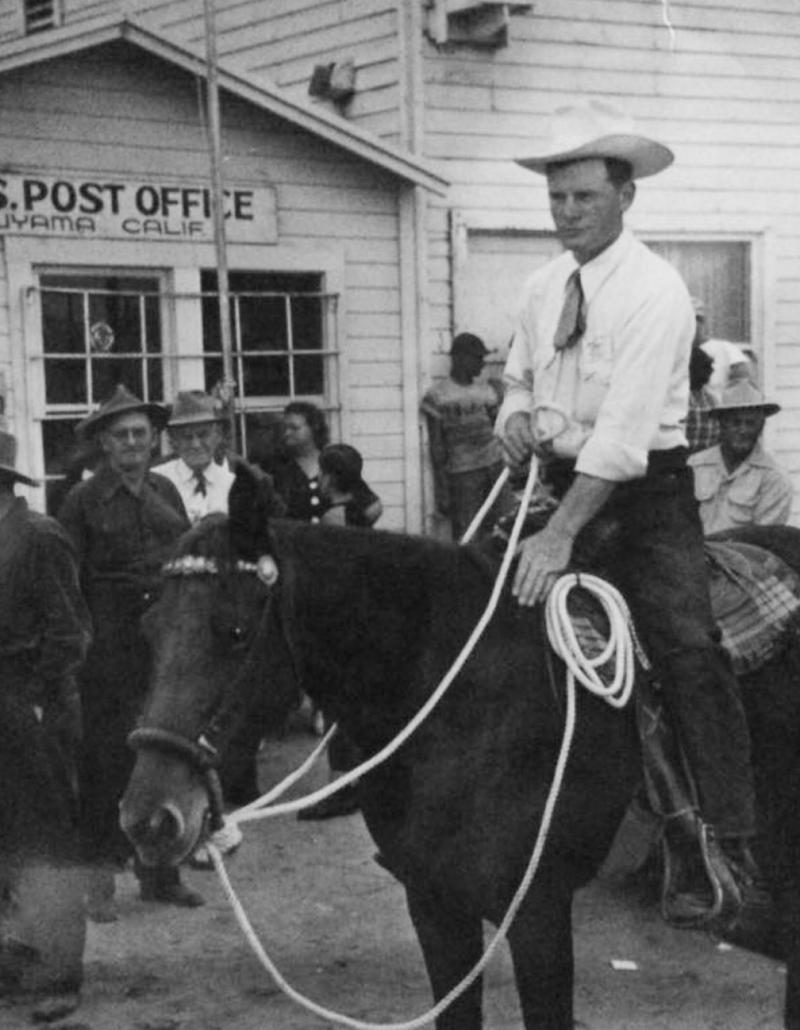 A cowboy on horseback with a lasso outside the New Cuyama post office, a crowd behind him.