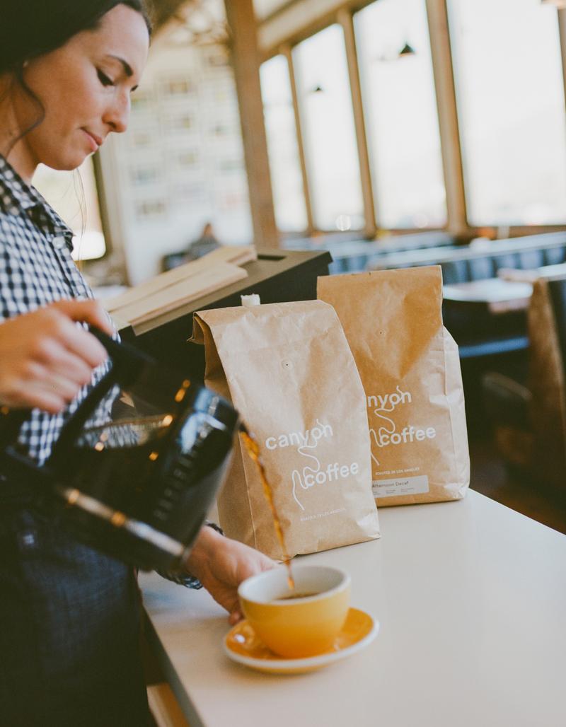 A barista pouring filter coffee into a yellow cup beside bags of Canyon Coffee.