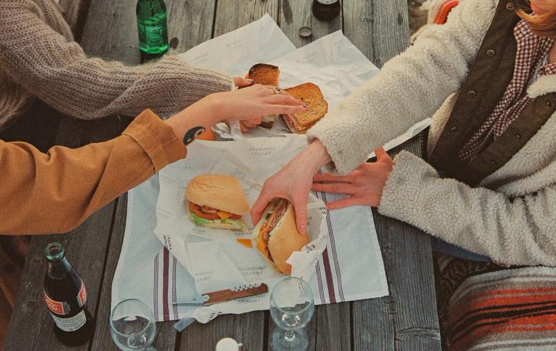 A group of friends picking up sandwiches on a picnic table