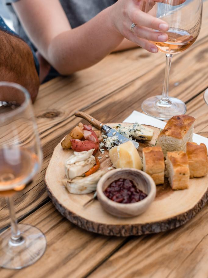 Hand reaching for rosé beside a cheese and bread board on a wood plank
