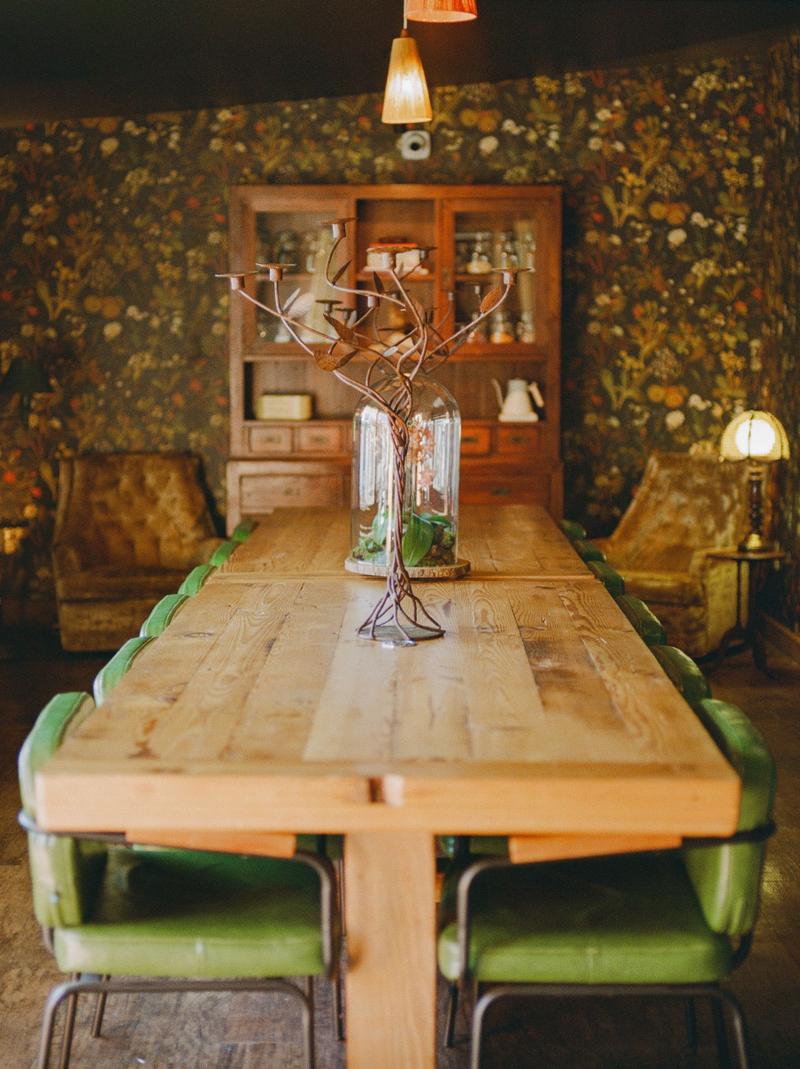A rustic dining table with green leather chairs against floral wallpaper