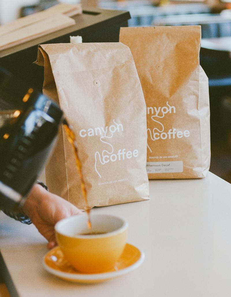 Hand pouring coffee into a mug beside two kraft bags labeled Canyon Coffee