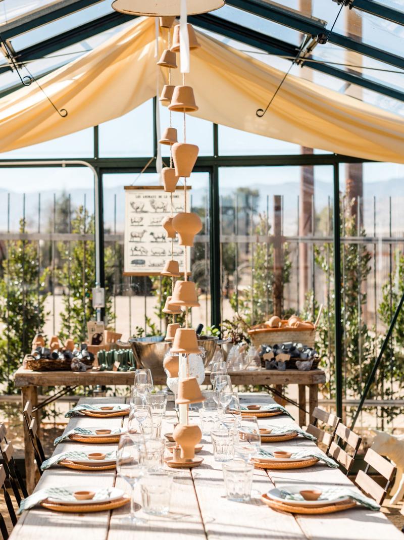 A table set under a glass greenhouse roof hung with a terracotta pot garland