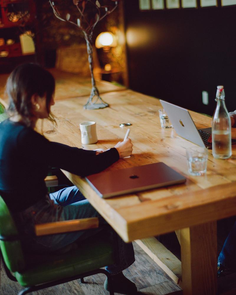 A guest working on a laptop at a wooden table by the window, a bottle of water and a vase of flowers beside them.