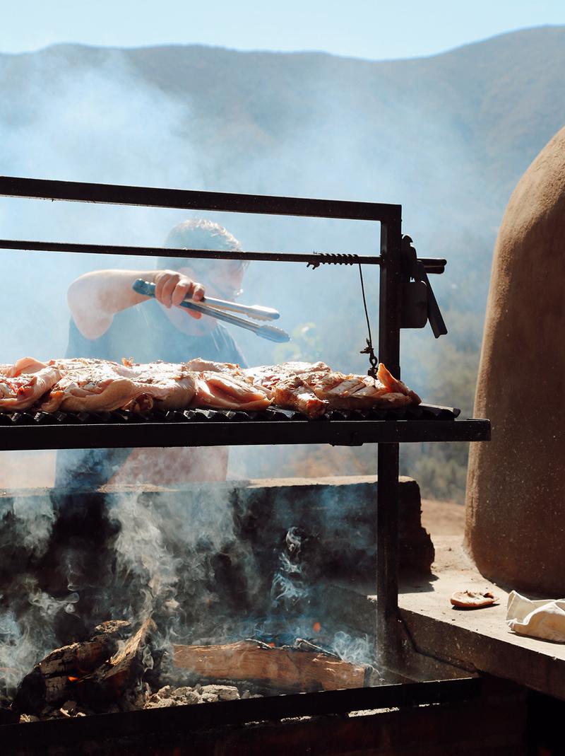 A cook tends chicken on an open-fire grill with mountains in the background