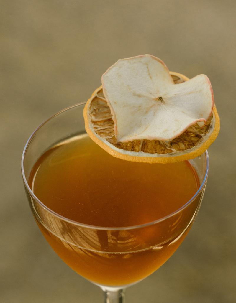 An amber cocktail in a stemmed glass, garnished with dried citrus and apple slices.