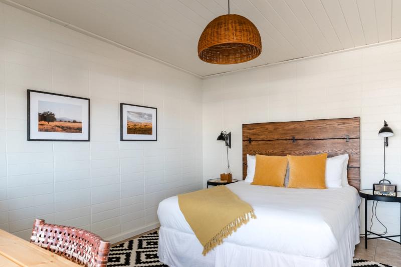 Motel room with woven pendant light, framed landscape prints and a mustard throw on the bed