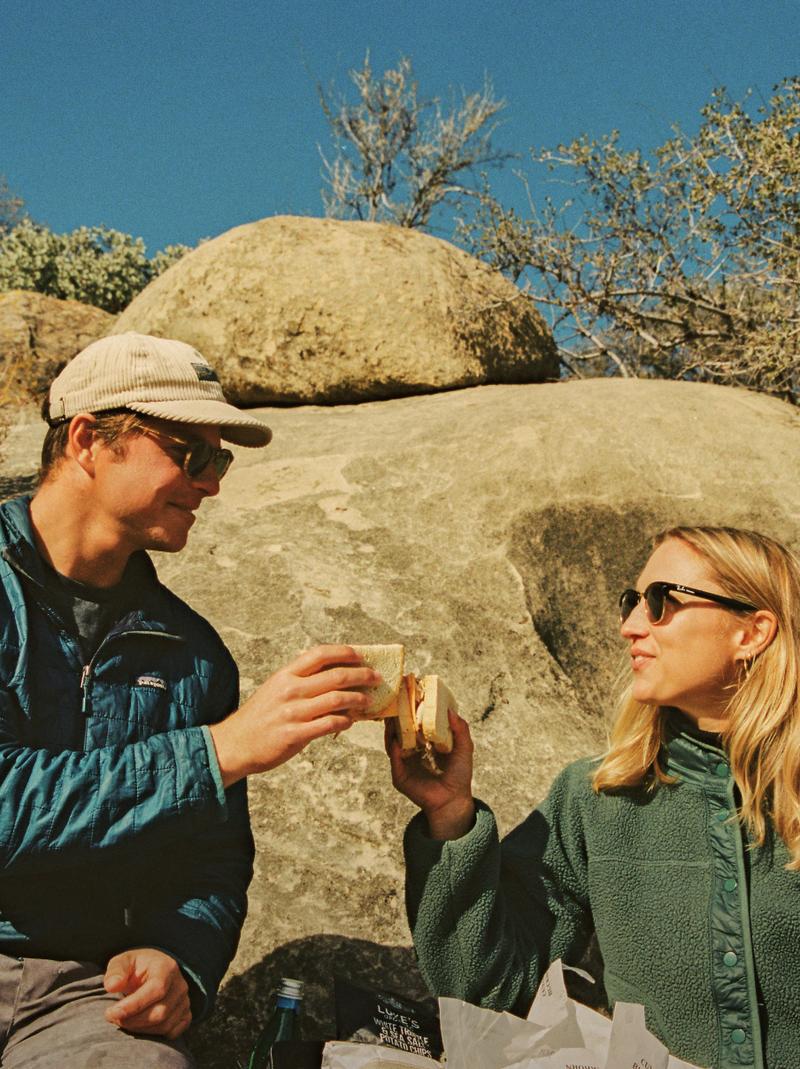 Two guests toasting with enamel mugs on a picnic among the sandstone boulders.