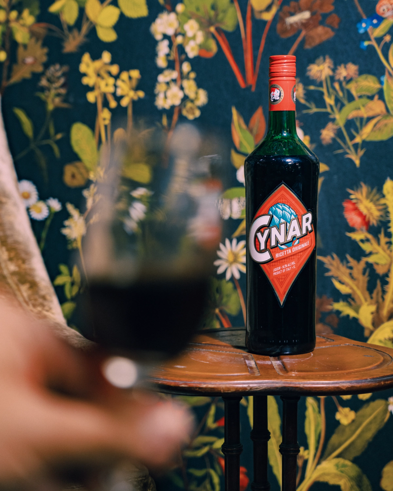 A bottle of Cynar amaro beside a glass, against floral wallpaper