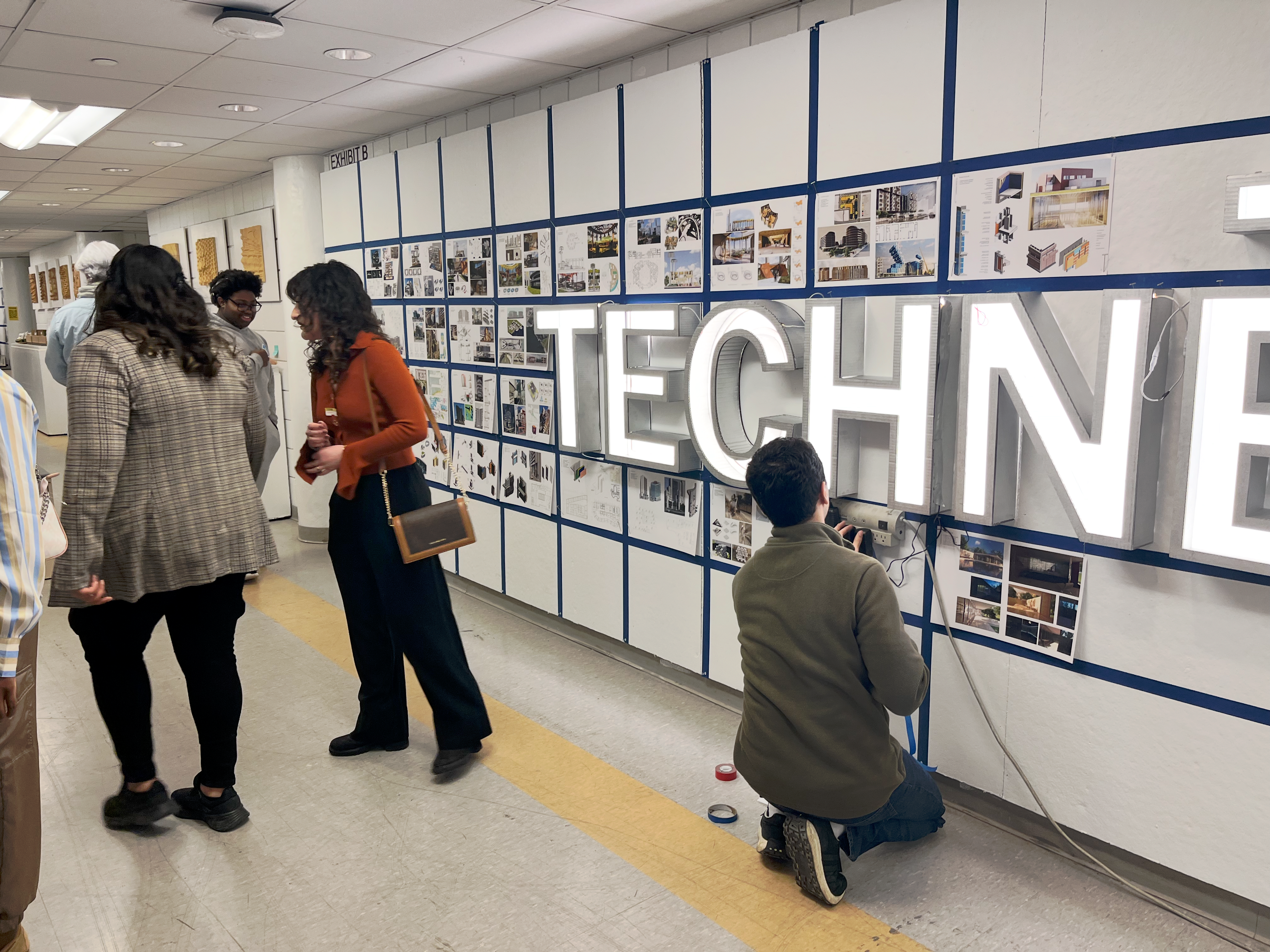 Students and visitors view architecture student work displayed on a wall grid during the TECHNE exhibition at CityTech, while a student kneels to install large illuminated letters spelling “TECHNE” in front of the display.