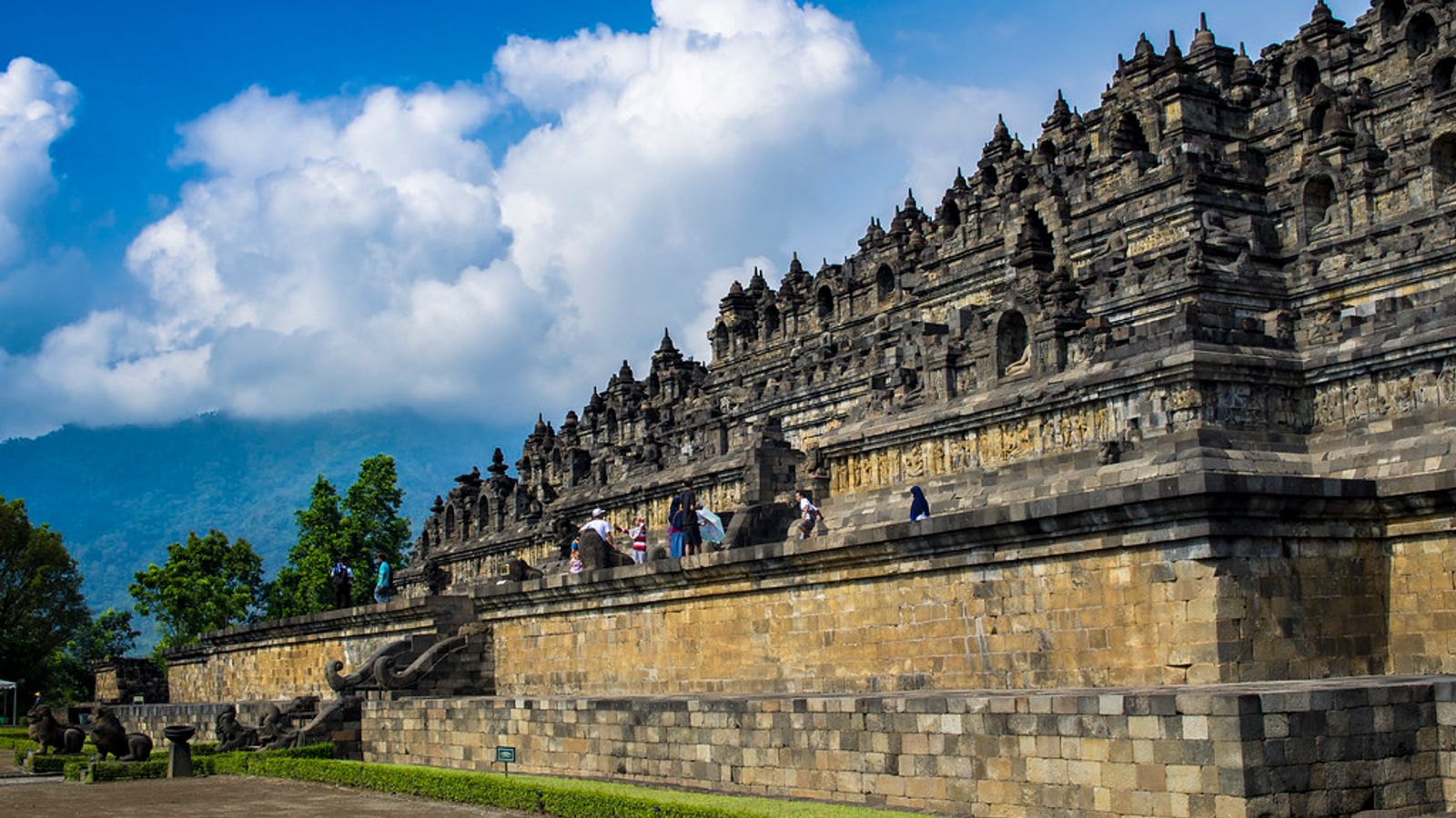 Visitors walking the stone terraces of Borobudur temple under a blue sky in Central Java