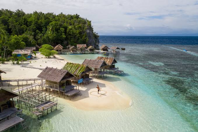 Papuan homestay bungalows on stilts over a shallow turquoise reef in Raja Ampat, with a white sandbar and jungle-covered karst behind