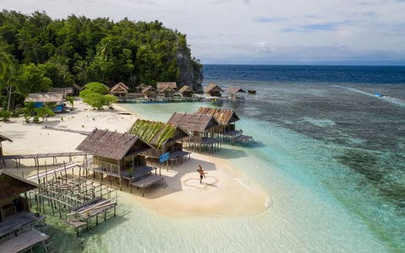 Papuan homestay bungalows on stilts over a shallow turquoise reef in Raja Ampat, with a white sandbar and jungle-covered karst behind