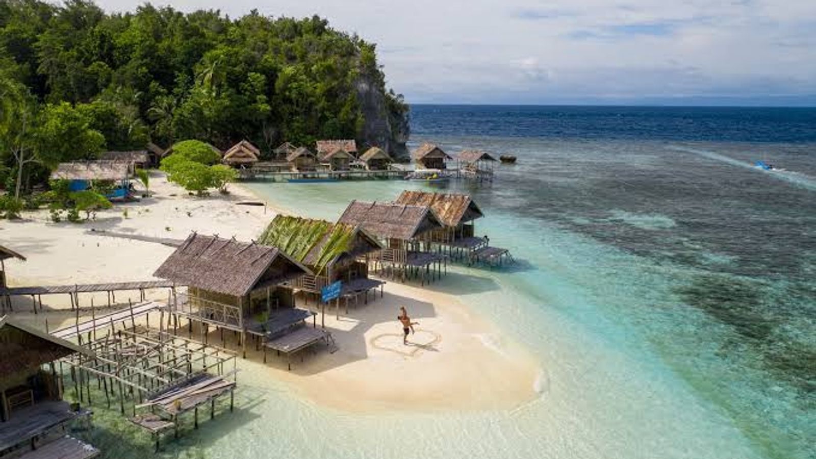 Papuan homestay bungalows on stilts over a shallow turquoise reef in Raja Ampat, with a white sandbar and jungle-covered karst behind