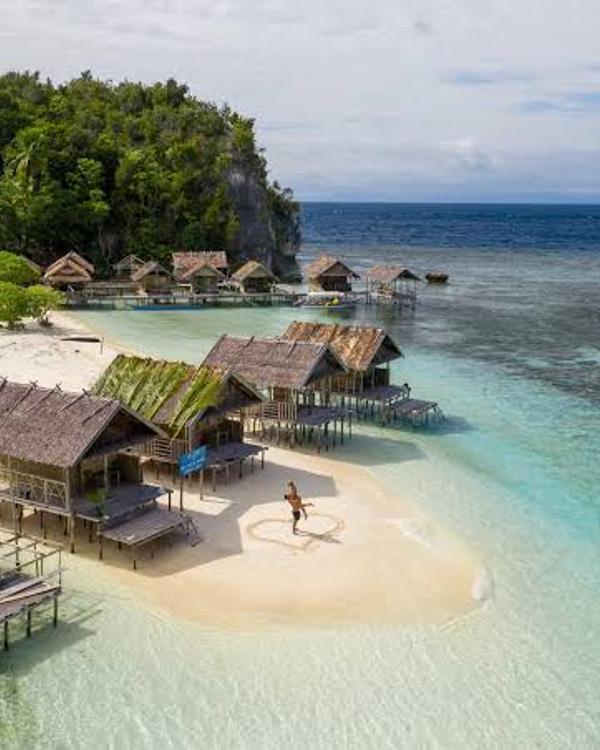 Papuan homestay bungalows on stilts over a shallow turquoise reef in Raja Ampat, with a white sandbar and jungle-covered karst behind