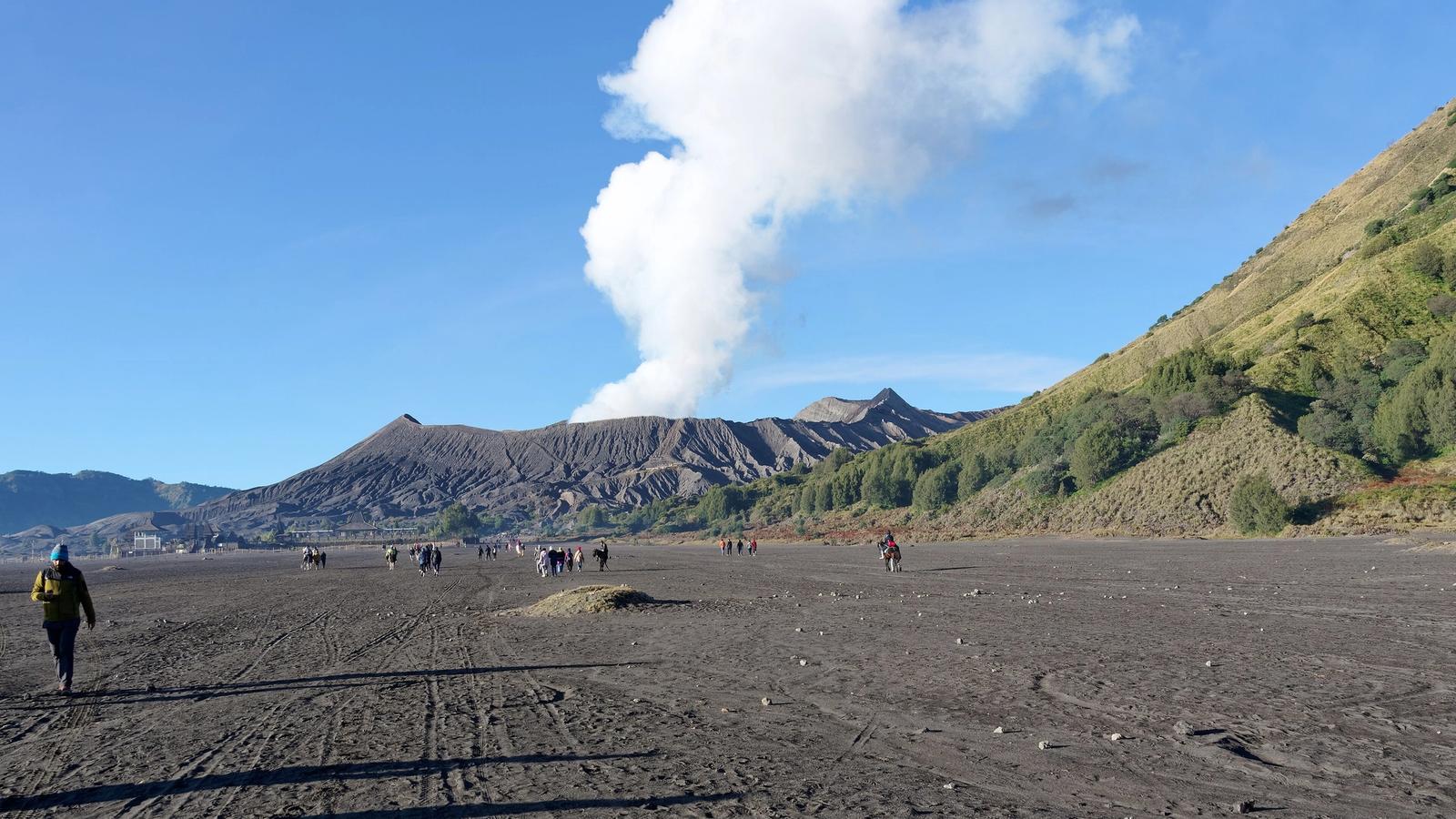 Visitors walking across the sea of sand towards the steaming crater of Mount Bromo on a clear morning in East Java