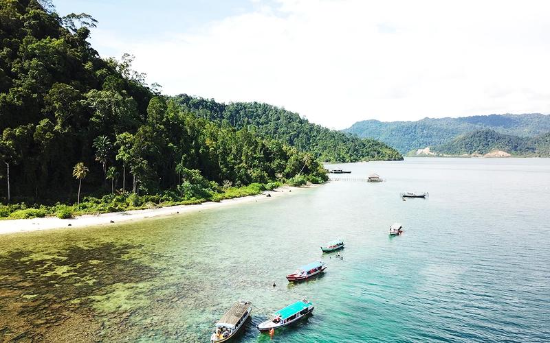 Wooden boats moored over shallow reef below a forested headland in Mandeh Bay, West Sumatra