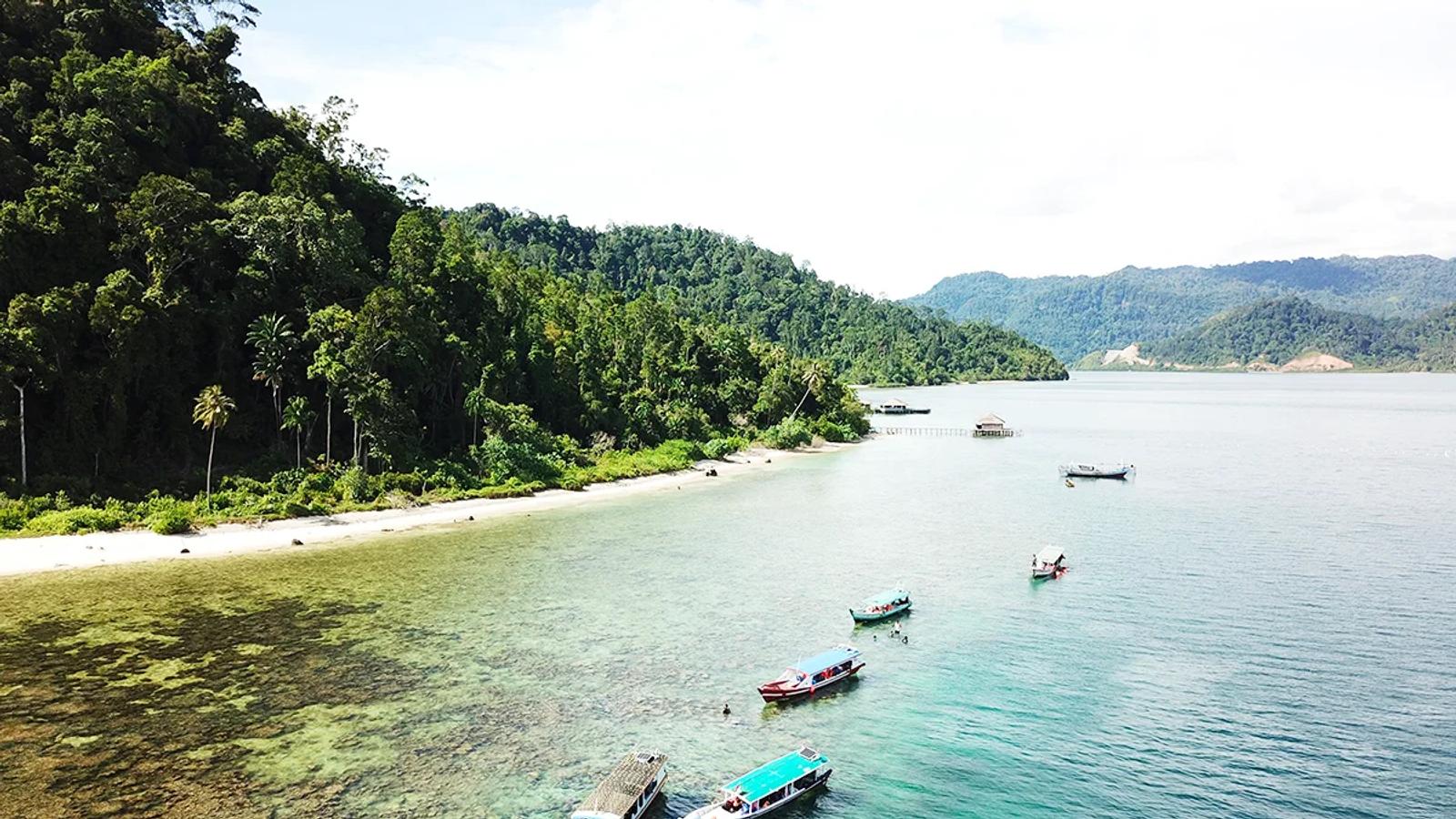 Wooden boats moored over shallow reef below a forested headland in Mandeh Bay, West Sumatra