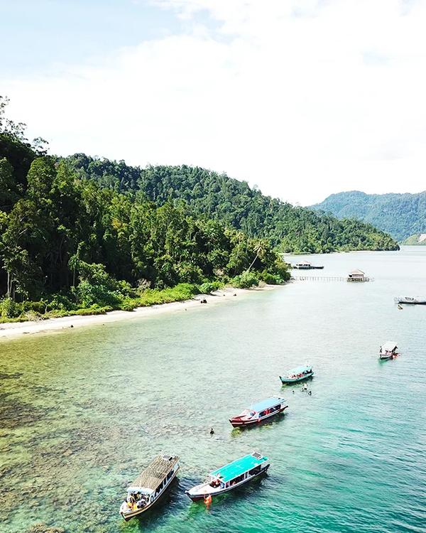 Wooden boats moored over shallow reef below a forested headland in Mandeh Bay, West Sumatra
