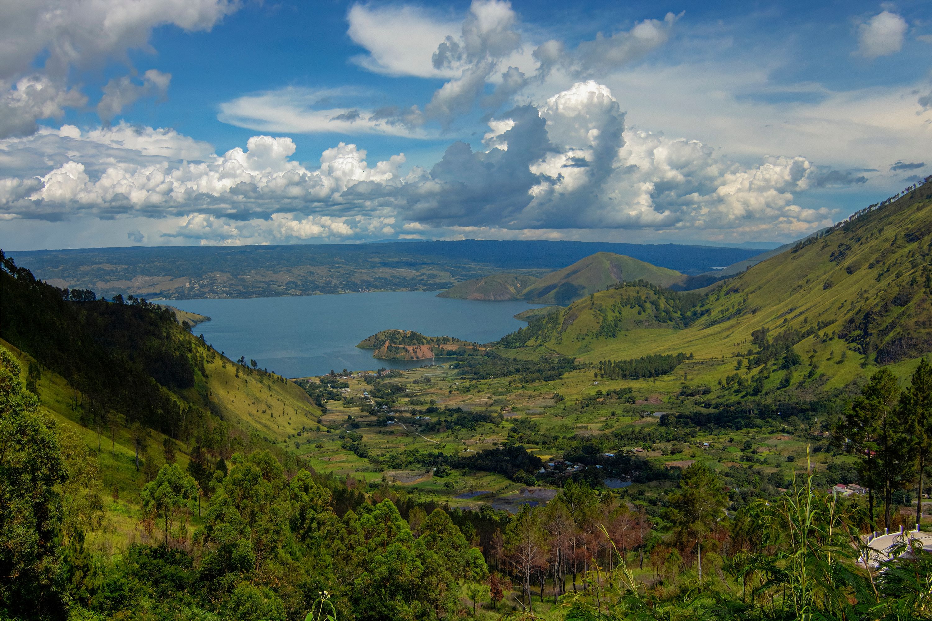 The green caldera rim of Lake Toba dropping to the water in North Sumatra, with a cultivated valley floor below and cumulus cloud over the far shore