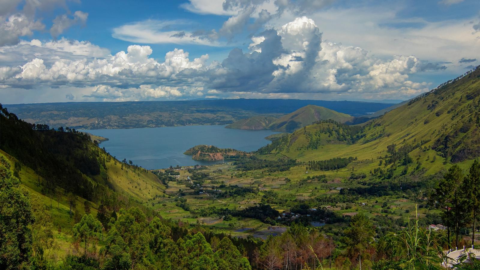 The green caldera rim of Lake Toba dropping to the water in North Sumatra, with a cultivated valley floor below and cumulus cloud over the far shore