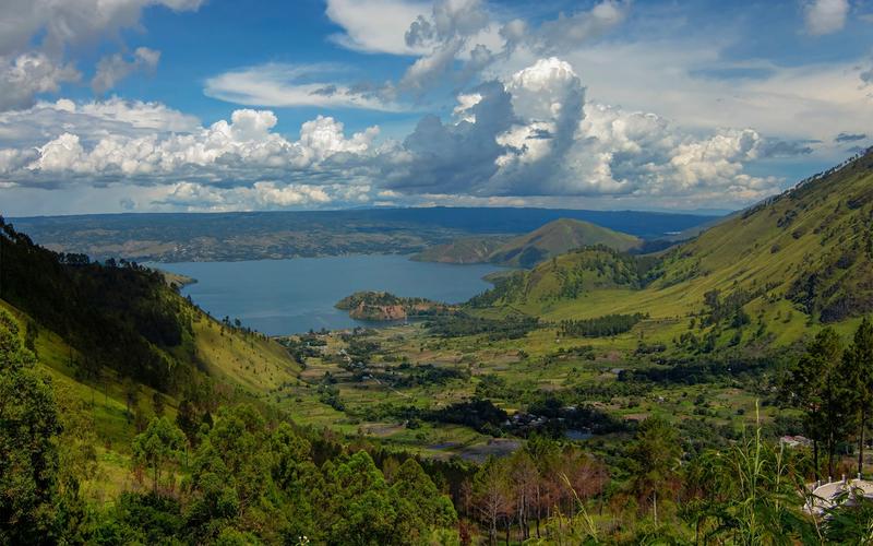 The green caldera rim of Lake Toba dropping to the water in North Sumatra, with a cultivated valley floor below and cumulus cloud over the far shore